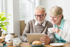 Ein Seniorenpaar sitzt am Frühstückstisch und schaut lächelnd auf ein Tablet