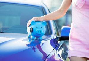 Ein Frau im rosa Business Kleid steht neben einem blauen Auto, auf dem ein Sparschwein steht