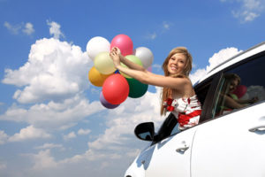 Eine lachende Frau lehnt sich aus einem Autofenster und hält dabei viele bunte Luftballons in den Händen