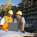 Zwei Männer mit Bauhelmen stehen vor Großbaustelle und studieren Baupläne