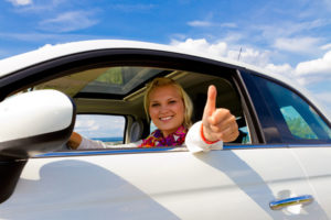 Eine junge Autofajhrerin lächelt und hält den erhobenen Daumen aus dem Autofenster