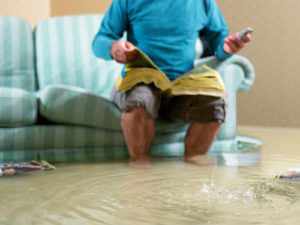 Ein Mann sitzt auf einem Sofa das im Hochwasser seiner Wohnung steht und sucht in den gelben Seiten nach einer Telefonnummer