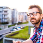 Junger Mann mit Bart und Brille steht auf einem Balkon und blickt erwartungsvoll in die Ferne