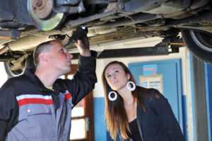 Eine junge Frau und ein Mechaniker unterscuhen ein Auto, das auf einer Hebebühne steht