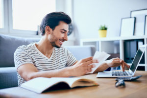 Ein lächelnder junger Mann sitzt am Couchtisch und beantragt über seinen Laptop einen Kredit