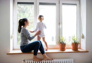Eine junge Mutter spielt mit ihrem Kind am Fenster und stellt ihre Füße auf die Heizung
