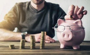 Ein junger Mann füttert ein Sparschwein mit Brille, neben dem ansteigende Münzstapel stehen