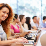 Eine lächelnde Studentin mit Laptop sitzt am Anfang einer Reihe Studenten im Hörsal
