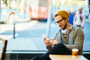 Ein junger Mann sitzt bei einem Kaffe, hat Kopfhörer um den Hals gelegt und tippt auf seinem Handy